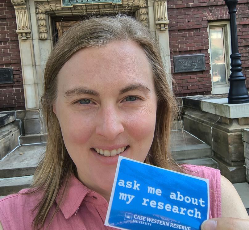 CWRU researcher Lydia Kisley posing for a selfie holding a sticker that reads, “ask me about my research.