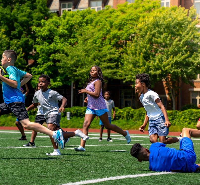 Participants in the National Youth Sports Program take part in a game of football at DiSanto Stadium on a sunny day.