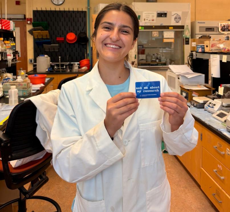 Fourth-year undergraduate student, Emilia Sanz-Rios, posing and holding a sticker that reads, “Ask me about my research.”