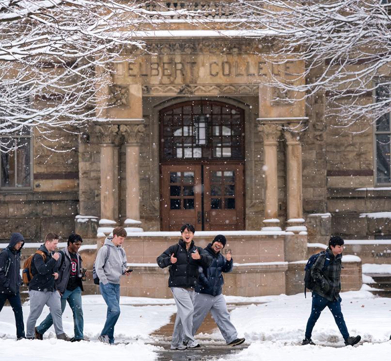 Students walking past Adelbert Hall in the snow, posing with thumbs up.