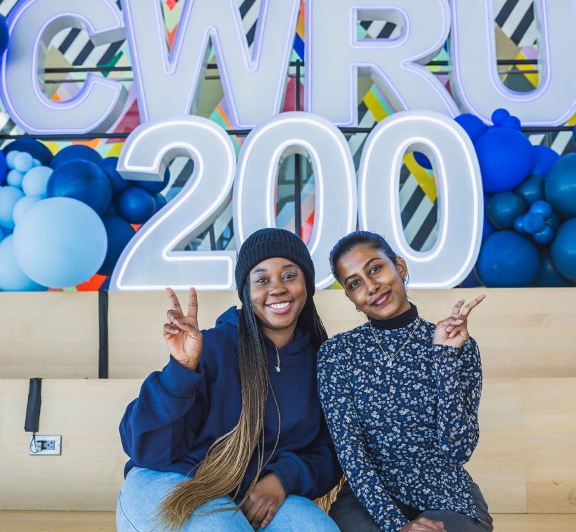 Two women posing with peace sign hand gestures in Tinkham Veale University Center in front of a balloon and letter display reading, “CWRU 200.”