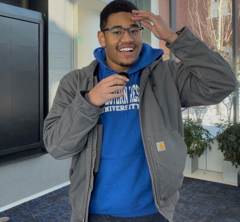 A student wearing a blue hoodie and glasses holding a small microphone in one hand, with his other hand on his forehead.