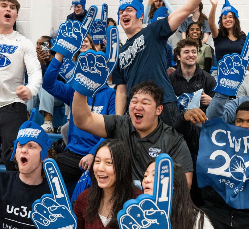 A large group of CWRU students wearing blue foam Spartan helmets and blue foam fingers, cheering on a basketball game at Horsburgh Gymnasium.