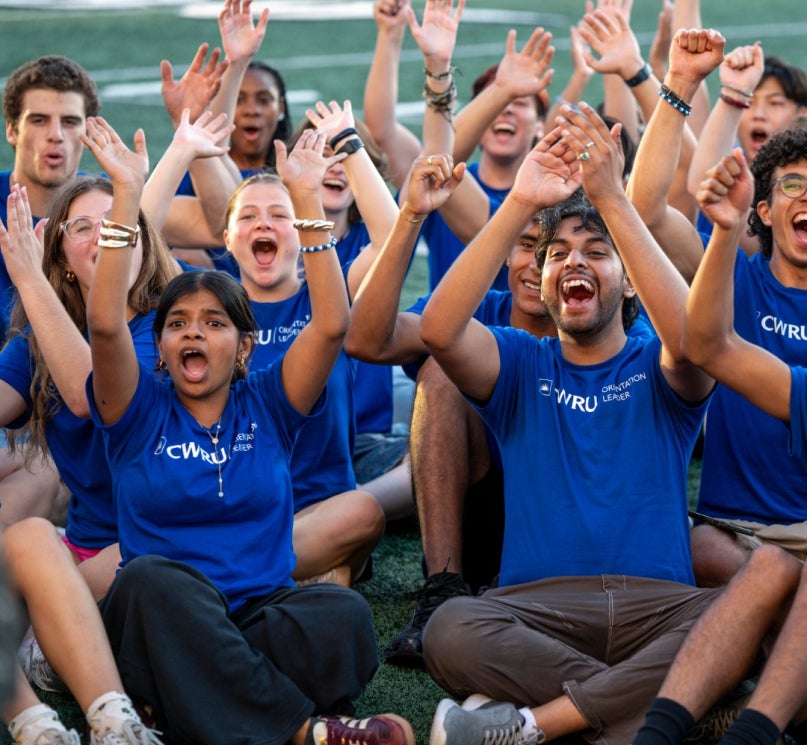 A group of CWRU orientation leaders sitting together on DiSanto Field in matching shirts, smiling and cheering.