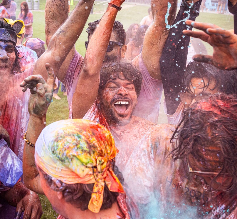 A group of students laugh and splash water on each other while covered in orange, yellow, purple and blue powder at the on-campus Holi celebration.