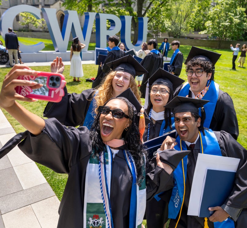 A group of five students in caps and gowns posing for a selfie in front of the “CWRU” sign on campus.