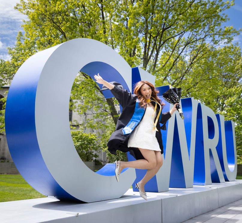 A student jumping in a cap and gown in front of the “CWRU” sign on campus.