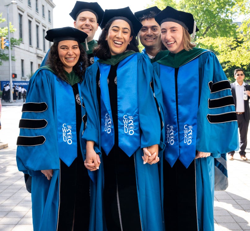 Five CWRU School of Medicine students pose for a photo in their academic regalia holding hands.