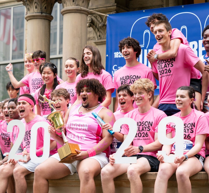 A group of first-year students wearing pink “Hudson Relays 2026” shirts pose outside Adelbert Hall, cheering and holding a trophy alongside white cutout numbers reading “2029.”