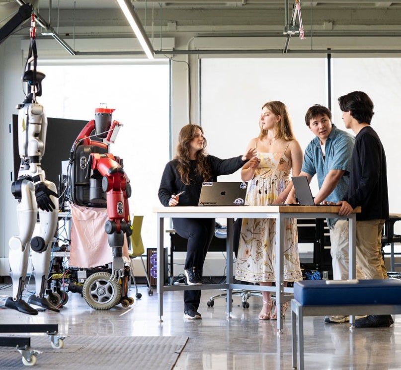 A group of six CWRU students talking in a collaboration space with two robots nearby.