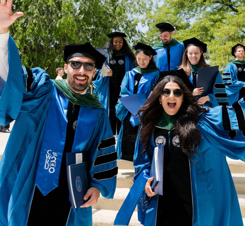 Two CWRU School of Medicine students waving to the camera with fellow graduates visible in the background.