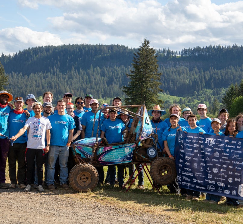 A group of CWRU Baja SAE members pose behind their competition vehicle, holding a sign featuring sponsor logos, in Oregon.