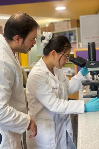 A scientist observes a student in a lab coat viewing a slide through a microscope. 