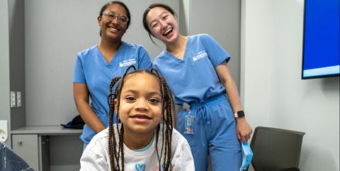 A young girl with braids in a white shirt poses in front of two dental students.