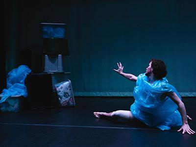 Photo of Joel Linebach wearing a plastic bag-like material dancing on the ground with an arm outstretched toward a pile of plastic material