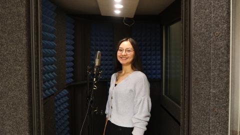 A woman stands smiling in a soundproof recording booth with blue acoustic panels, next to a microphone.