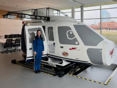 Amanda Rowe stands in front of a white helicopter simulator wearing a blue jumpsuit.
