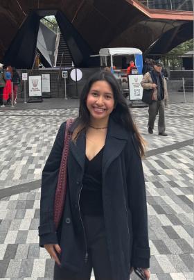 Zelan Eroz Espanto stands in a plaza wearing a black coat with people walking behind her.