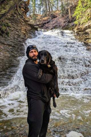 A man dressed in black holds a black labrador retriever in front of a small waterfall.