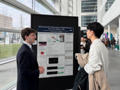 A student in a suit talks to another in a white shirt holding a tan coat in front of a black board with a poster on it.