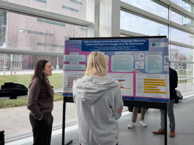 Two people stand in front of a medical research poster in front of a glass wall.