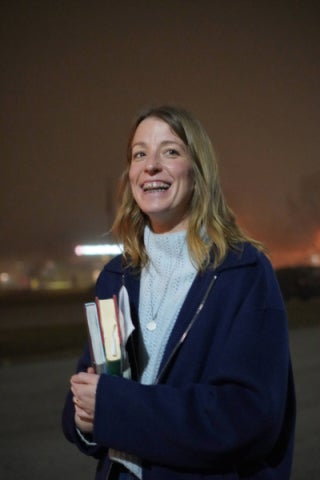 Elizabeth Ginsburg stands smiling in a black cardigan holding a stack of books in front of a dusk sky.