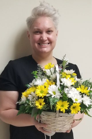 Raquel Dauch smiles while posing in dark blue scrubs holding a basket bouquet of white and yellow flowers.