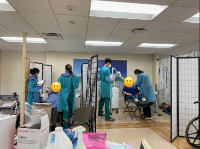 Dental students in green gowns work on two dental patients divided by screens.