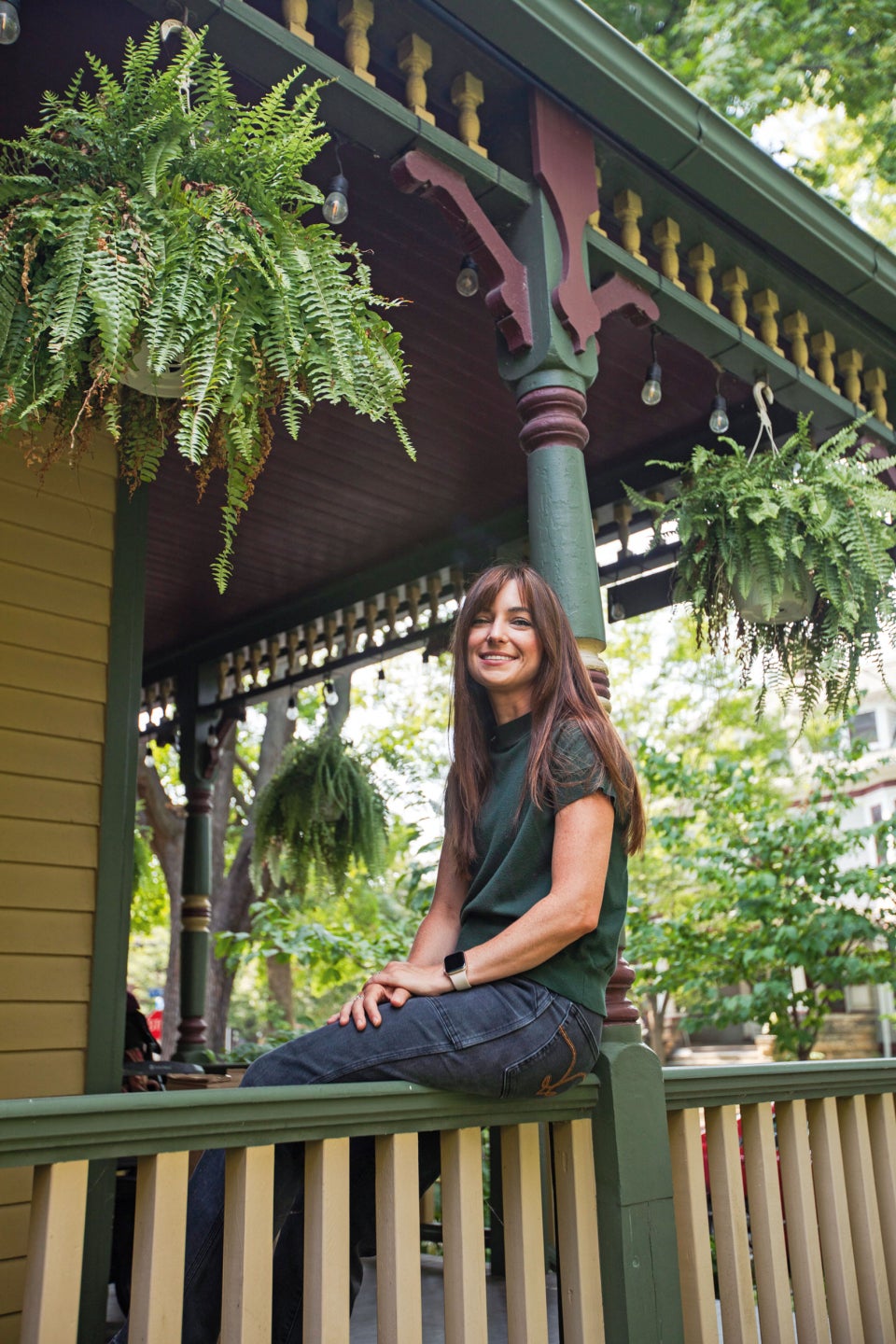 A photograph of Marissa Scavuzzo sitting on a porch railing. Lush greenery and hanging ferns surround her in the background.