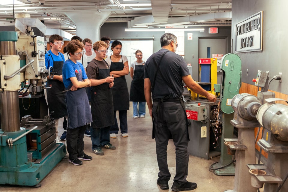 A group of students wearing safety goggles and black aprons attentively observes an instructor operating a machine.