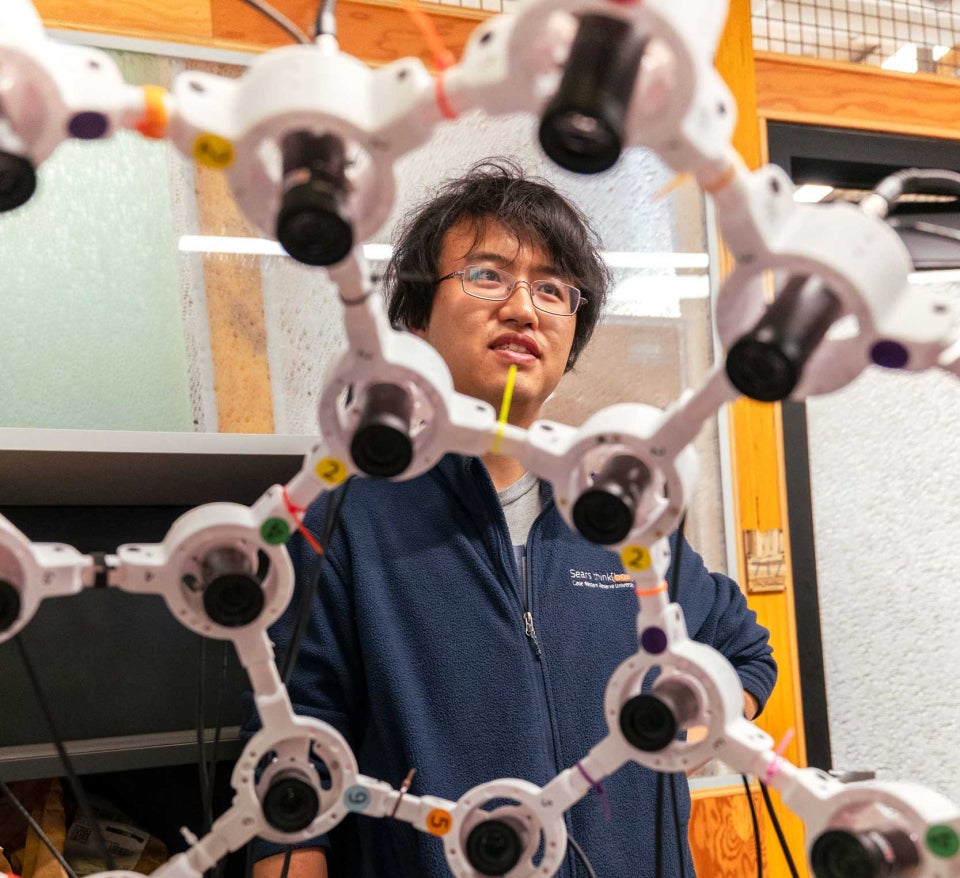 A person in a Sears think [box] jacket stands behind a geometric frame mounted with cameras. 