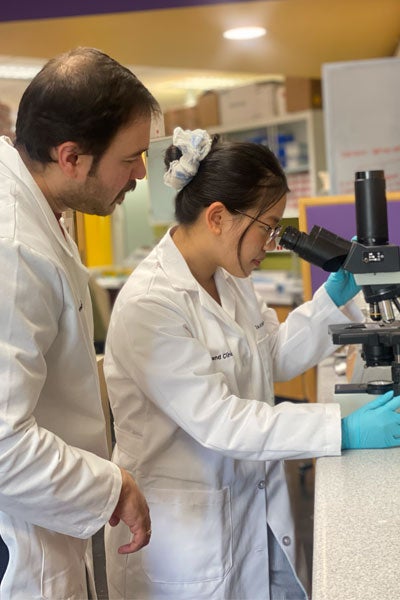 A scientist observes a student in a lab coat viewing a slide through a microscope. 