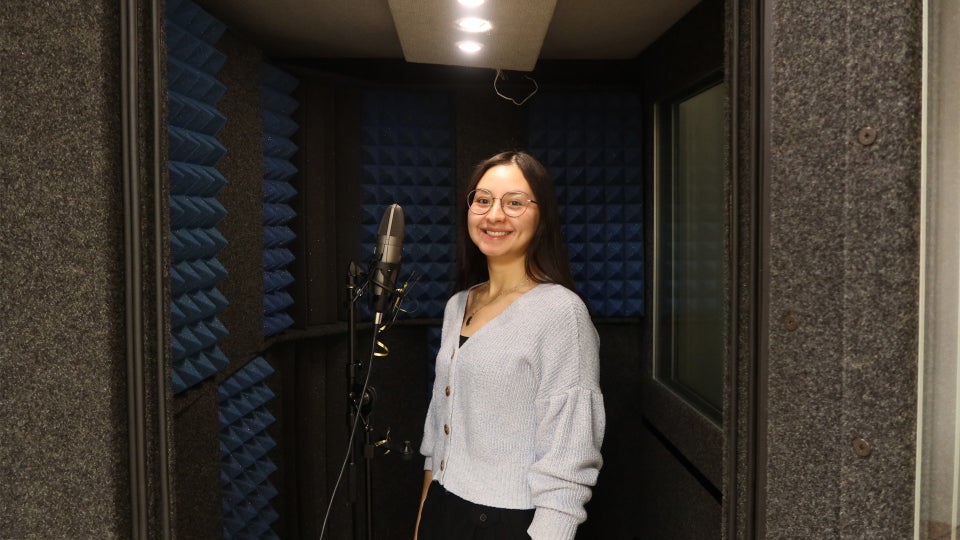 A woman stands smiling in a soundproof recording booth with blue acoustic panels, next to a microphone.