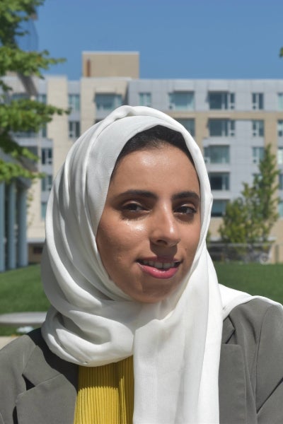Hind Alsabti poses in a white head scarf and grey jacket in front of a building on a sunny day with blue skies above