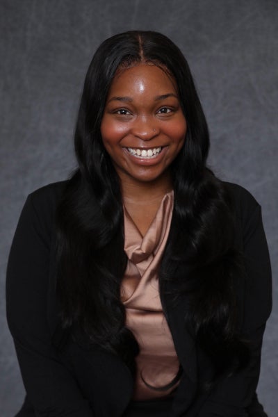 Miriam Ashford in pink blouse and black jacket poses in front of grey background