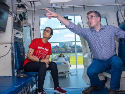 A woman in a red shirt and a man in a plaid button down sit inside a helicopter flight simulator.