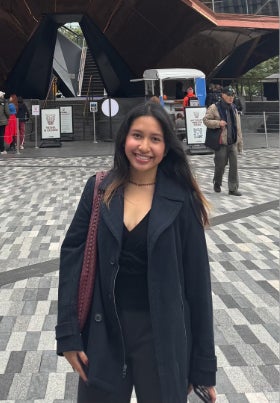 Zelan Eroz Espanto stands in a plaza wearing a black coat with people walking behind her.