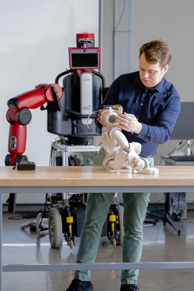 Austin stands at a worktable in a robotics lab, adjusting the head of a small white robot seated on the table.