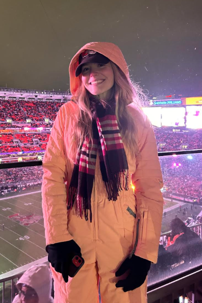 Caroline Deyoe stands smiling in a brown coat in Browns stadium with crowds of people in the stands behind her