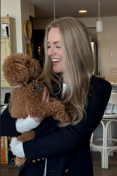 A blond woman in a black jacket smiles at the small red curly-haired dog she is holding.