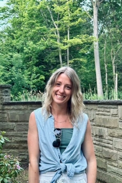 Elizabeth Ginsburg stands smiling in a blue shirt in front of a stone wall with green trees behind.