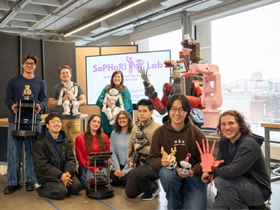 Nine members of the SaPHaRI Lab pose together in a robotics lab, smiling at the camera while holding various robots and robotic devices