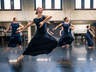 A main dancer and two dancers behind her all leap in a studio wearing blue dresses. Three dancers can be seen practicing separately behind them. 