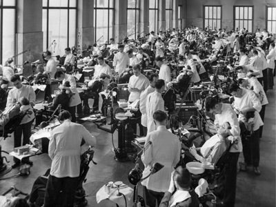 A large, bustling dental clinic with rows of dentists in white coats attending to patients in chairs. The scene is busy and clinical with focused activity.