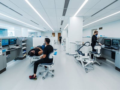 A modern dental clinic with sleek white interiors and several patients receiving care. Dentists and staff, wearing black uniforms and masks, attend to patients in dental chairs.