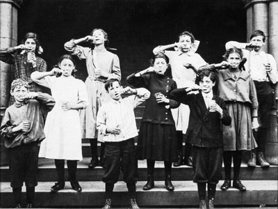 A black and white photo shows a group of children standing in two rows on steps, holding glasses. They humorously make funny faces, conveying playfulness.