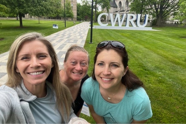 Three women stand in front of a grassy lawn and pathway with CWRU statue behind them.