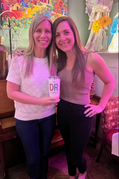 Two women pose together while holding a mug that says "it's DR actually"