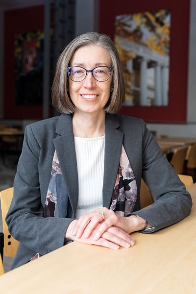 A woman with short gray hair, glasses, and a gray blazer smiles while sitting at a table in a warmly lit room. 