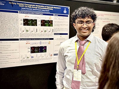 Photo of Kanishk Neerumalla smiling as he discusses his research in front of his poster at NCUR